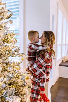 a woman in plaid pajamas hugging her son near a christmas tree