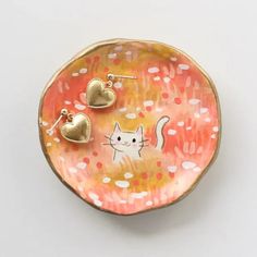 two gold heart shaped earrings on top of a pink and yellow plate with a cat design