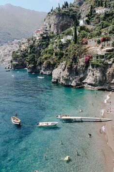 people are swimming in the water near boats on the beach and mountains behind them,