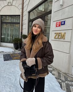 a woman standing in front of a building with snow on the ground and holding onto her purse