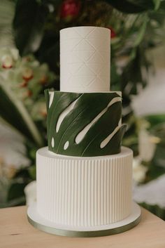 a three tiered white and green wedding cake with greenery in the back ground