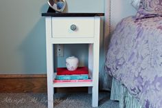 a small white nightstand next to a bed with a purple bedspread on it