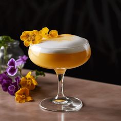 a glass filled with liquid sitting on top of a wooden table next to purple and yellow flowers