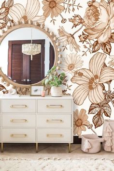 a white dresser sitting next to a mirror on top of a wooden cabinet in front of a floral wallpaper