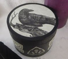 a black bird sitting on top of a wooden box next to a purple cup and pen