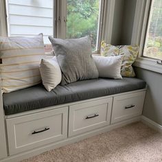 a window seat with several pillows on it in front of two windows, one is white and the other is gray