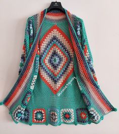 a crocheted sweater hanging on a hanger next to a white wall with an orange, blue and green pattern