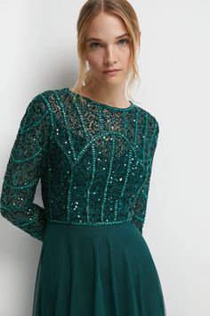 a woman wearing a green dress with sequins on the top and sleeves, standing in front of a white wall