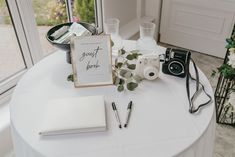 a white table topped with a camera, notebook and other items next to a window