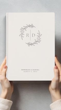 two hands holding up a white book with the initials on it and an elegant wreath