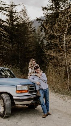 a man and woman standing in front of a truck on a dirt road near trees