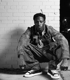 a black and white photo of a young man sitting on the ground in front of a brick wall