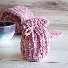 two crocheted bags sitting next to a bowl on a wooden table with a cup in the background