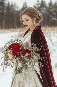 a woman in a white dress holding a bouquet and red velvet coat standing in the snow