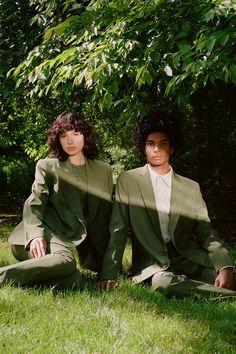 two people sitting on the grass in front of some trees and one person is wearing a suit