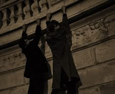 two men standing next to each other on the side of a building with their hands in the air