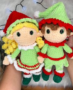 two crocheted elves are holding onto each other's stuffed toys, with christmas lights in the background