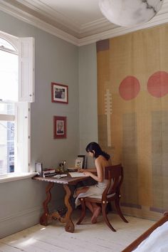 a woman sitting at a desk in front of a window with a painting on the wall behind her