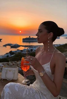 a woman sitting on a bench holding a wine glass in front of the ocean at sunset