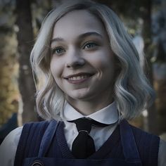 a woman with blonde hair and blue overalls smiles at the camera while standing in front of trees