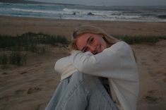 a woman is sitting on the beach with her arms over her head and looking at the camera