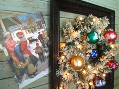 a christmas tree with ornaments and lights in front of a painting on a wooden wall