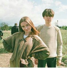 two young people standing next to each other in a field with one person wearing a brown jacket