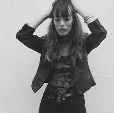 a woman with her hands on her head standing in front of a white wall wearing jeans and a jacket