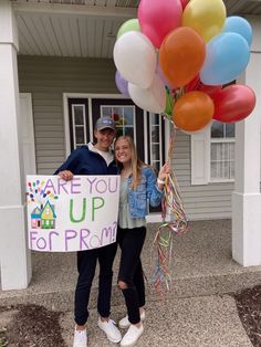 a man and woman are holding balloons in front of a house that says are you up for prom?