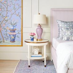 a white bed sitting next to a pink table with flowers on it and a painting behind it