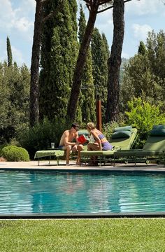 two people sitting at the edge of a swimming pool with lounge chairs and trees in the background