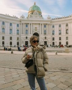 a woman standing in front of a large building with a cup of coffee on her hand