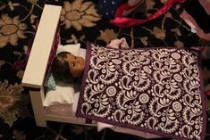 a doll in a box sitting on the floor next to other dolls and accessories,