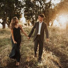 a man and woman holding hands while walking through the grass with trees in the background