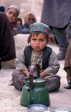 a little boy sitting on the ground with a tea pot in front of him and other people around him