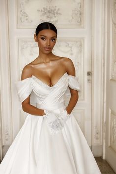 a woman in a white wedding dress standing next to a wall and wearing an off the shoulder bow