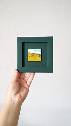 a hand holding up a small green frame with a painting on the inside of it