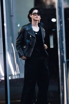 a woman standing in front of a glass door talking on a cell phone while wearing black pants and a leather jacket