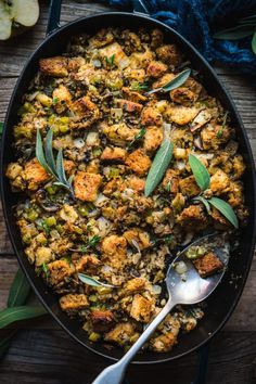 a pan filled with stuffing and sage leaves on top of a wooden table next to an apple