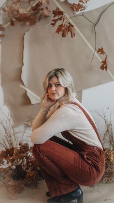 a woman squatting on the ground in front of a wall with leaves and branches