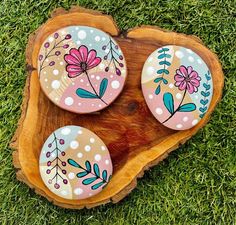 three painted rocks sitting on top of a piece of wood in the grass with flowers