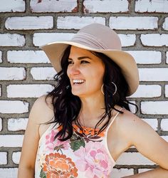 a woman wearing a hat standing in front of a brick wall with her hands on her hips