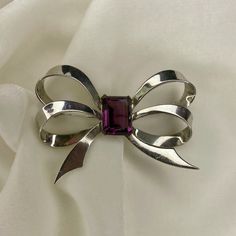 I received this vintage unsigned brooch in a lot of estate jewelry. It is a minimalist silver tone bow shape, with a central purple rhinestone. It is a nice size, measuring 2.5 inches long. There was a missing rhinestones, otherwise the brooch was in good condition.  The pin mechanism is in good working order.   This is an elegant statement piece which will dress up any outfit. Vintage Brooch With Decorative Bow For Evening, Vintage Brooches With Decorative Bow For Evening, Vintage Evening Brooch With Decorative Bow, Silver Brooch With Decorative Bow, Vintage Bow Brooch For Formal Occasions, Vintage Evening Brooches With Decorative Bow, Silver Brooches With Decorative Bow, Vintage Formal Brooch With Bow, Vintage Jewelry With Decorative Bow For Formal Occasions