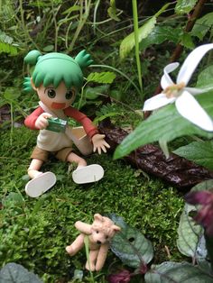 a small doll sitting on the ground next to some plants and flowers, with another toy in the foreground