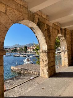 an arched stone wall overlooks a harbor with boats on the water and mountains in the distance