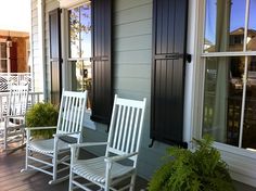 two white rocking chairs sitting on a porch next to windows with shutters and potted plants