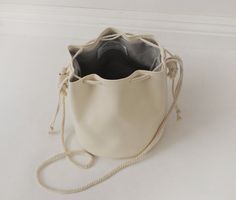 "This unique bucket bag features soft, off-white faux (fake) leather, cotton rope shoulder strap, and a drawstring closure. You can tie the cotton rope to wear this bucket bag long or short. Details: -Dimensions when laid flat: 9\" length (top to bottom), 7.5\" diameter round bottom -Shoulder strap measures approx. 45-50\" total -Soft, off-white faux (fake) leather -Solid light color interior lining -No internal pocket Because each bag is made by hand, the measurement is approximate and may vary Casual Cream Bucket Bag Tote, Cream Bucket Bag With Adjustable Strap For Shopping, Cream Casual Bucket Bag For Shopping, Casual Cream Bucket Bag For Daily Use, Beige Bucket Bag With Adjustable Strap, Cream Leather Bucket Bag With Braided Handles, Cream Bucket Bag For Everyday Use, Cream Bucket Bag For Everyday Spring Use, Casual Cream Bucket Bag For Everyday