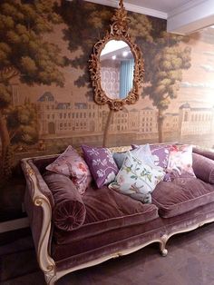 a couch with many pillows on it in front of a wallpapered background and mirror