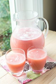 two glasses and a pitcher filled with pink liquid sit on a table next to each other