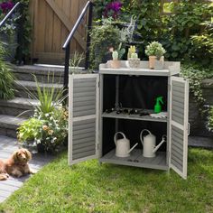 a dog laying in the grass next to an outdoor storage cabinet with plants and watering cans